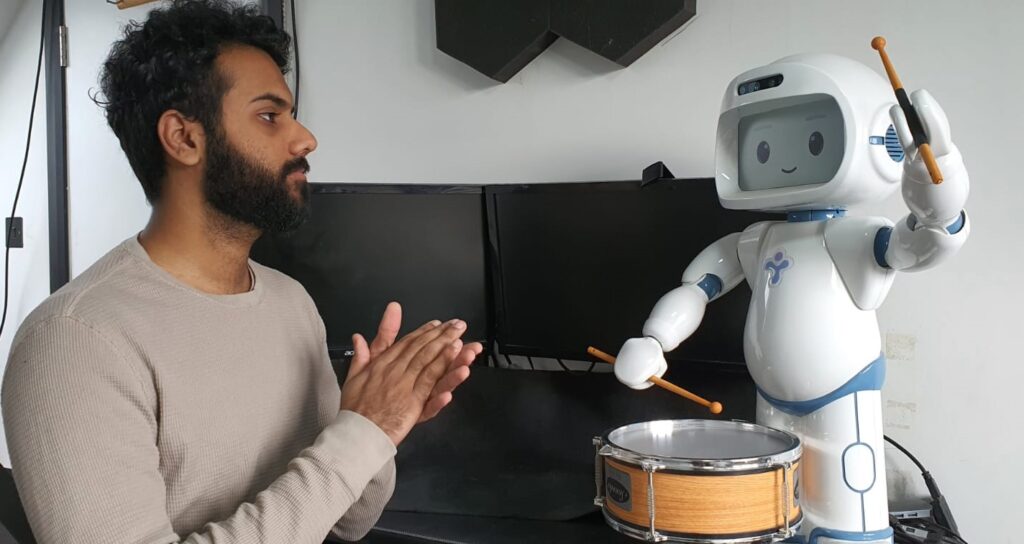 Humanoid robot playing drum alongside man clapping hands