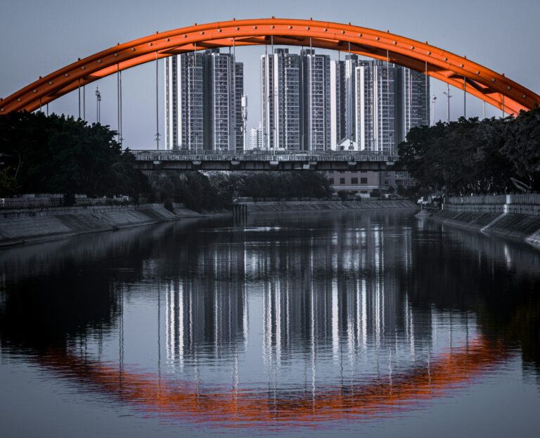 Photograph of a steel arch bridge spanning an urban waterway, with tower blocks in the background. The bridge is reflected in the water below.